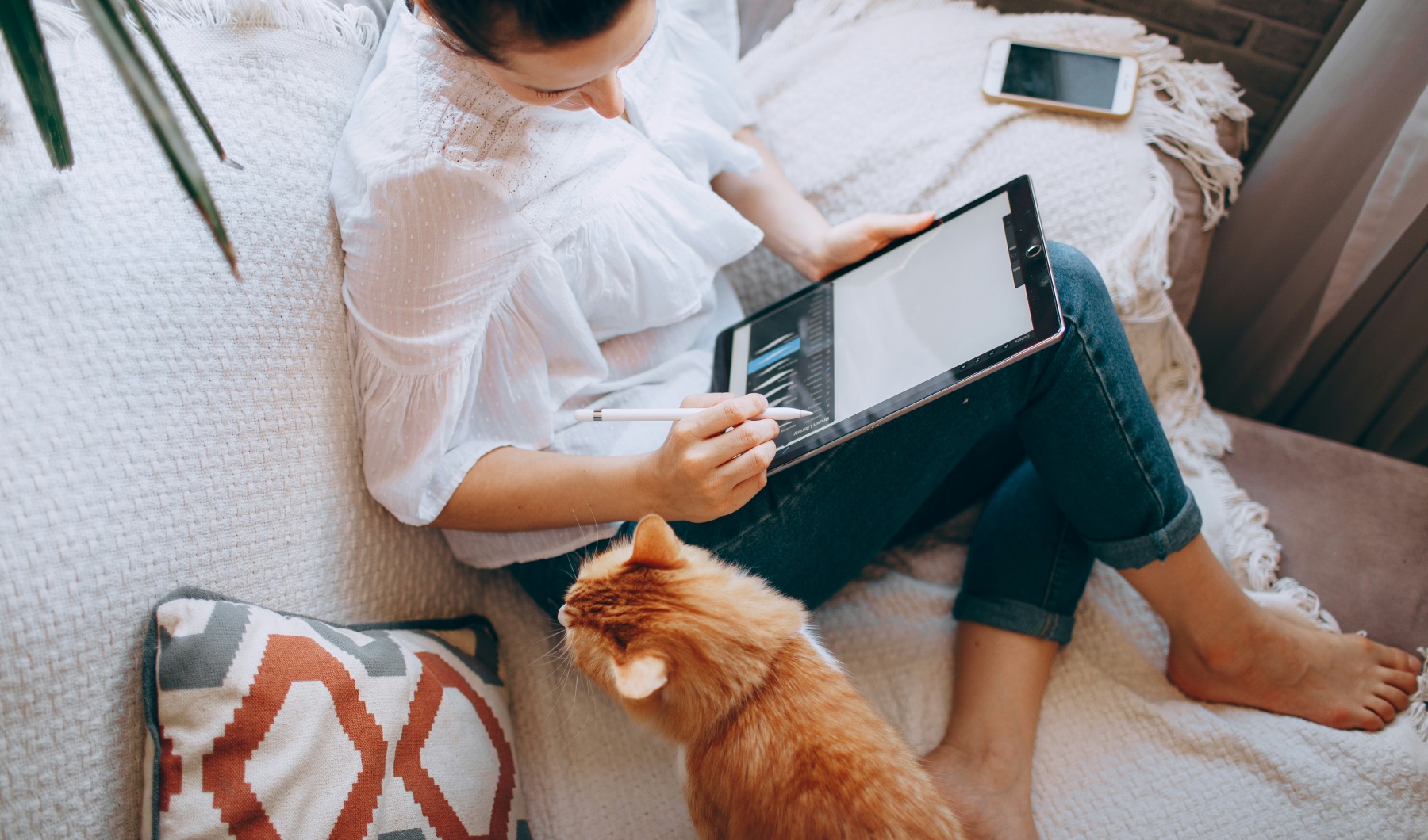 Woman sits on couch with cat while drawing on a tablet device.